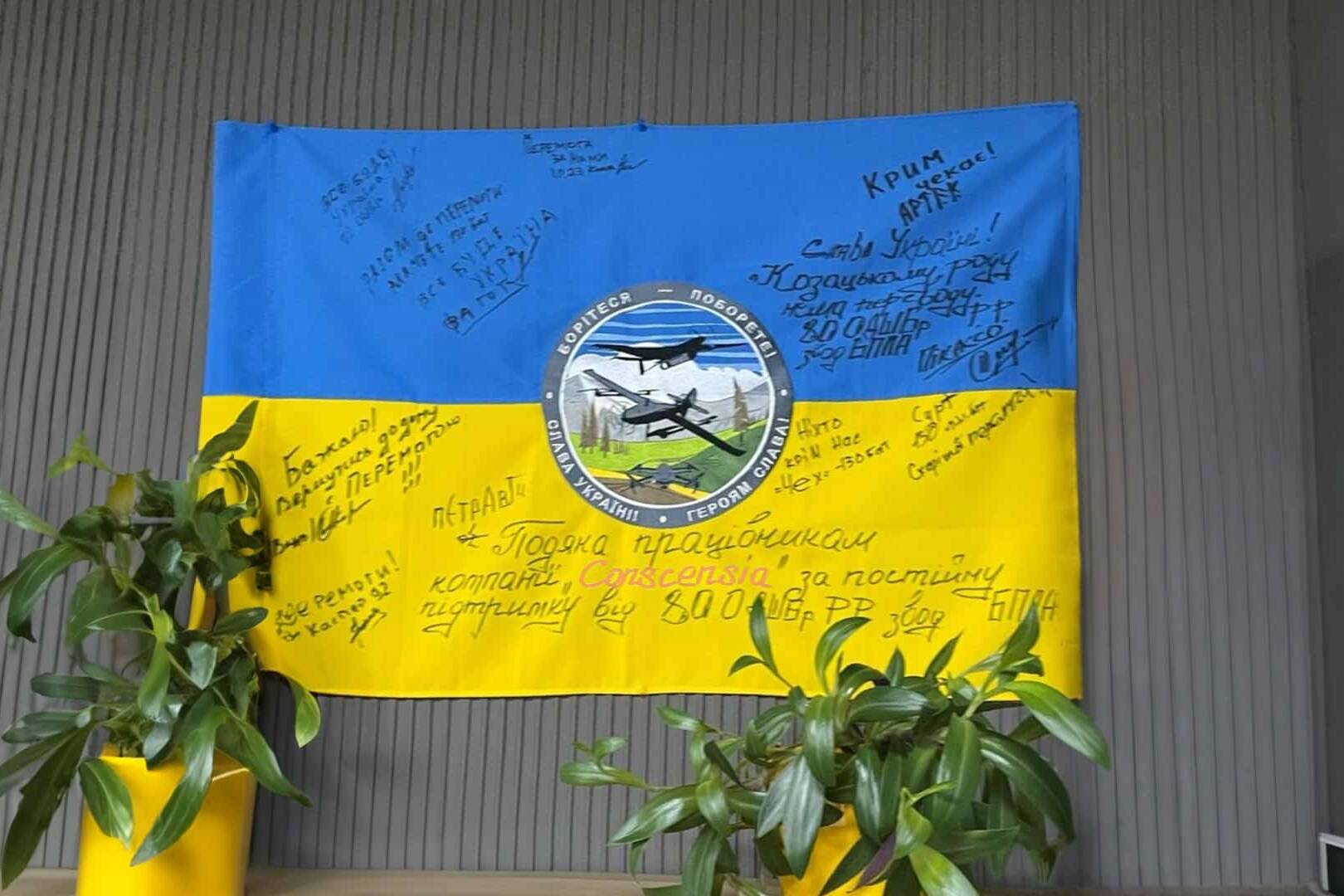 Et blåt og gult ukrainsk flag med signaturer og en besked på ukrainsk, der udtrykker taknemmelighed til Conscensias ansatte for deres støtte, hængende på en væg.